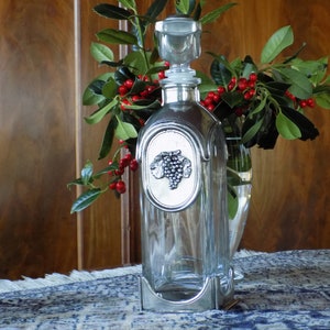 Puede incluir: Una decantador de vidrio transparente con una base y un tapón de metal plateado. La decantador presenta un diseño de vid en la parte delantera. La decantador está sentada sobre un mantel estampado azul y blanco.