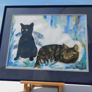 May include: A framed watercolor painting of two cats. One cat is black and sitting, the other is tabby and lying down. The background is a blue and green abstract design.