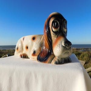 May include: A ceramic dog figurine in a lying down position. The dog has a white body with brown and blue spots, and a black, brown, and orange head. The figurine is on a white surface with a blue sky and ocean in the background.
