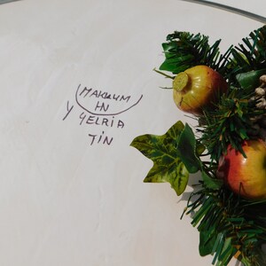 May include: A white plate with a handwritten message that says "MAKUM HN Y GELRIA TIN" and a green garland with red and yellow apples.
