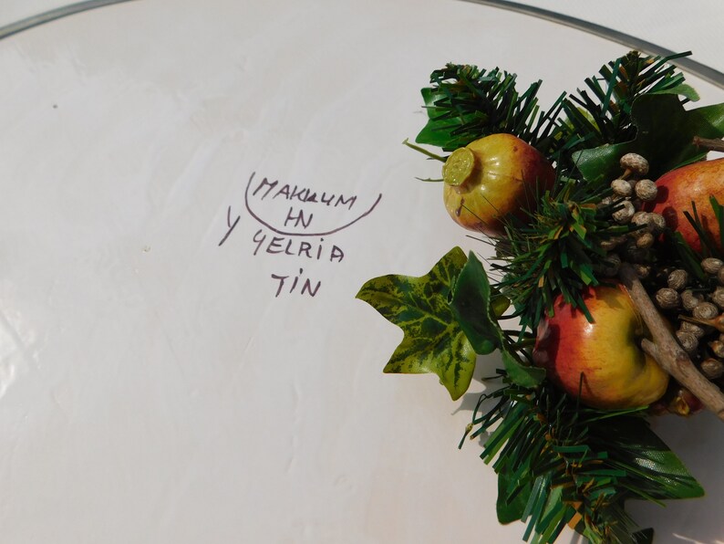 May include: A white plate with a handwritten message that says "MAKUM HN Y GELRIA TIN" and a green garland with red and yellow apples.