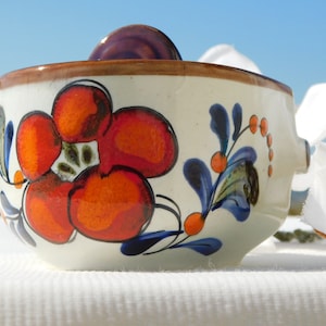 Op de afbeelding: Een witte keramische kom met een bruine rand en een bloemenmotief. De kom heeft een grote rode bloem met oranje accenten en blauwe en groene bladeren. De kom staat op een wit oppervlak.