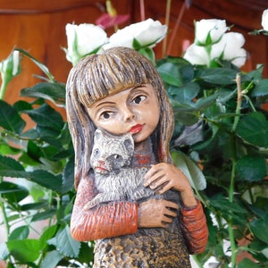 Puede incluir: Una figurita de cerámica de una niña con el pelo largo y castaño que sostiene un pequeño gato blanco. La figurita está rodeada de rosas blancas.