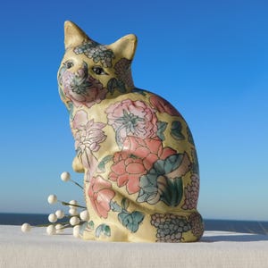 Puede incluir: Figura de gato de cerámica con un estampado floral en rosa, azul y verde sobre fondo amarillo. El gato está sentado con las orejas erguidas. Ramitas decorativas blancas frente al gato.