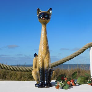 Op de afbeelding: Een lange keramische kattenfiguur met een slank lichaam en lange nek. De kat heeft een bruin lichaam met donkerblauwe accenten en blauwe ogen. Decoratieve patronen sieren het lichaam. De figuur staat op een wit oppervlak, met een touw en bladeren op de achtergrond.