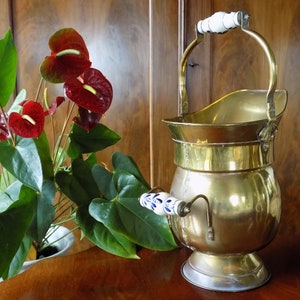 May include: A gold-colored metal coal scuttle with a white porcelain handle and a blue and white porcelain accent on the side. The scuttle is sitting on a wooden surface.