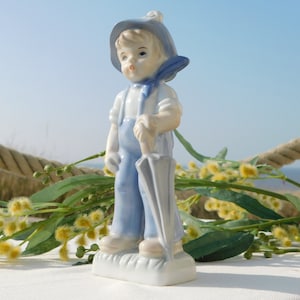 May include: A porcelain figurine of a boy wearing a blue hat and overalls, holding a blue umbrella. The figurine is standing on a white base and is surrounded by yellow flowers.