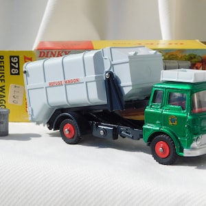 May include: A green and white Dinky Toys 978 Refuse Wagon with a tipping action. The toy truck has a white refuse bin with the words "Refuse Wagon" printed on the side. The box is yellow with red and black text.