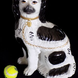 K&ouml;nnte beinhalten: Eine schwarze und wei&szlig;e Porzellan-Hundefigur mit einem goldenen Kettenhalsband. Der Hund sitzt mit leicht geneigtem Kopf. Die Figur steht auf einem schwarzen Hintergrund.