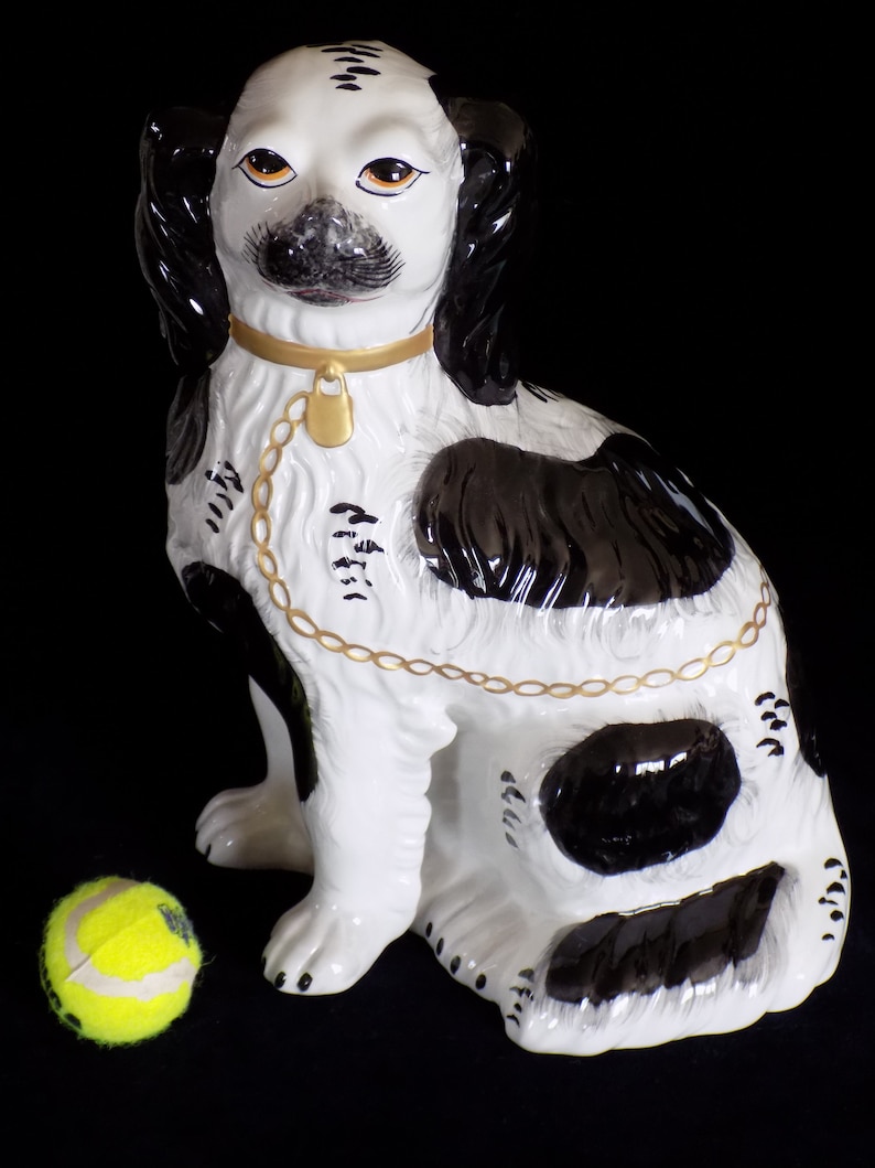K&ouml;nnte beinhalten: Eine schwarze und wei&szlig;e Porzellan-Hundefigur mit einem goldenen Kettenhalsband. Der Hund sitzt mit leicht geneigtem Kopf. Die Figur steht auf einem schwarzen Hintergrund.