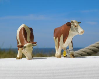 May include: Two toy cows with brown and white markings stand on a white surface against a blue sky. One cow is grazing, while the other stands upright. A rope is visible in the background, suggesting a nautical theme.