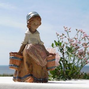 Może przedstawiać: Porcelanowa figurka dziewczyny w niebiesko-białym chustce i brązowej sukience, niosącej dwa wiadra. Figurka stoi na białym tle z bukietem różowych kwiatów w tle.