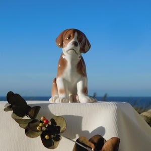 Op de afbeelding: Een keramieken hondenbeeldje, bruin en wit, in zittende positie. De hond heeft een serieuze uitdrukking, tegen een blauwe lucht en zee. Decoratief blad is op de voorgrond.