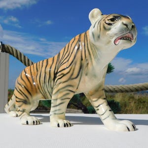 Könnte beinhalten: Eine Keramik-Tigerfigur mit orangefarbenen und schwarzen Streifen. Der Tiger steht auf einer weißen Oberfläche und hat den Mund offen.