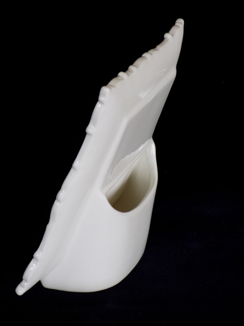 Może przedstawiać: Biała ceramiczna ramka na zdjęcia z falistym brzegiem i małym otworem z tyłu do trzymania zdjęcia. Ramka jest zaprojektowana do ustawiania w pozycji pionowej.