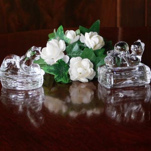 Puede incluir: Dos figuras de animales de cristal transparente, posiblemente gatos u nutrias, sobre una superficie de madera oscura. Flores blancas y hojas verdes se disponen entre las figuras. Las figuras de cristal son detalladas y reflejan la luz.