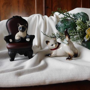 Op de afbeelding: Twee Siamese kattenbeeldjes. Eén zit in een miniatuur donkerbruine stoel, de andere poseert op een wit oppervlak. De katten hebben crèmekleurige lichamen, donkerbruine markeringen en blauwe ogen. Een rode halsband siert een kat.