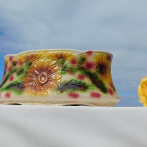 May include: A ceramic planter featuring a raised relief design of yellow sunflowers and green leaves against a cream-colored background with scattered pink spots. The bowl has a scalloped top edge and a wide, rounded base with a smooth finish.
