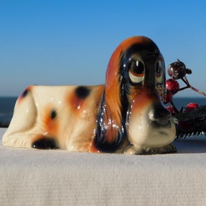 May include: A ceramic dog planter in a lying down position. The dog has a cream-colored body with black and orange spots, and a brown and black head. The planter is set against a blue sky and ocean background.