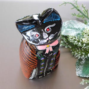 Puede incluir: Una figurita de gato de madera negra con detalles pintados en naranja, azul, rosa y blanco. El gato lleva una pajarita y tiene una expresión caprichosa.