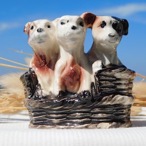 May include: A ceramic figurine of three white and brown dogs with black noses and eyes, sitting in a black and brown basket. The dogs are looking forward and the basket is made of woven material.