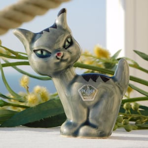 Puede incluir: Una figurita de cerámica gris de un gato con marcas negras y ojos azules. La figurita tiene un diseño estilizado y está de pie sobre una superficie blanca. El gato tiene una pequeña nariz rosa y una marca blanca en el pecho. La figurita tiene una pequeña etiqueta en la espalda con el texto "SYLCO" y una corona.