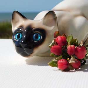 May include: A ceramic Siamese cat figurine with a cream-colored body, dark brown face mask, and bright blue eyes. The cat is positioned in a crouching pose next to a small cluster of red berries with green leaves, featuring a glossy finish.