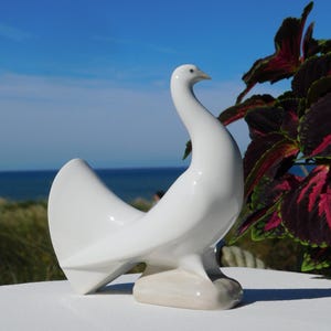 May include: A white ceramic dove figurine with a sleek, modern design. The bird has a curved neck and stylized wings, perched on a light beige base. The background features a blue sky, ocean, and green foliage.