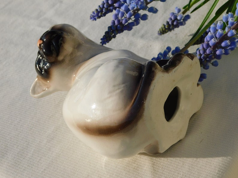 May include: A ceramic figurine of a Siamese cat in a sitting position. The cat is white with brown markings and has blue eyes. The figurine is glazed and has a glossy finish.