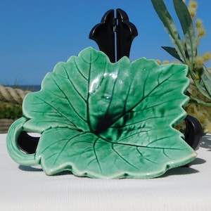 Könnte beinhalten: Eine grüne, keramikartige, blattförmige Schale mit detaillierten Adern, die auf einem schwarzen Ständer ruht. Die Schale hat eine glänzende Oberfläche. Der Hintergrund zeigt einen blauen Himmel und ein Seil.