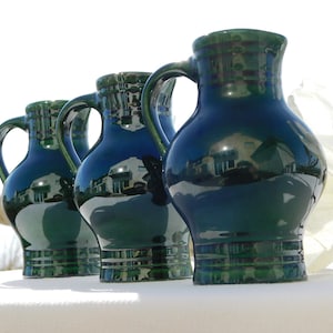 Op de afbeelding: Drie groene keramische kannen met handvatten, elk van een andere grootte. De kannen zijn geglazuurd en reflecteren de omgeving.