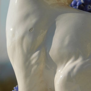 May include: A white ceramic sculpture of a woman's torso with a close-up view of the back. The sculpture is smooth and has a slight sheen. The woman's back is arched and her shoulders are rounded. The sculpture is lit from the side, casting shadows across the surface.
