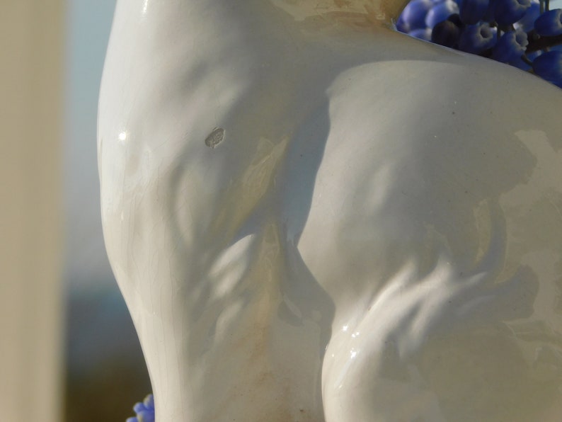 May include: A white ceramic sculpture of a woman's torso with a close-up view of the back. The sculpture is smooth and has a slight sheen. The woman's back is arched and her shoulders are rounded. The sculpture is lit from the side, casting shadows across the surface.