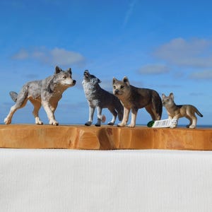 May include: Four wolf figurines of varying sizes standing on a natural-edged wooden slab. The wolves are depicted in shades of gray, brown, and tan. One wolf is shown howling with its head tilted upward, while the others stand in different poses.