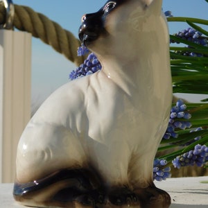 May include: A ceramic figurine of a Siamese cat sitting with its head turned to the side. The cat is white with black markings and has blue eyes. The figurine is sitting on a white surface with a bouquet of purple flowers in the background.