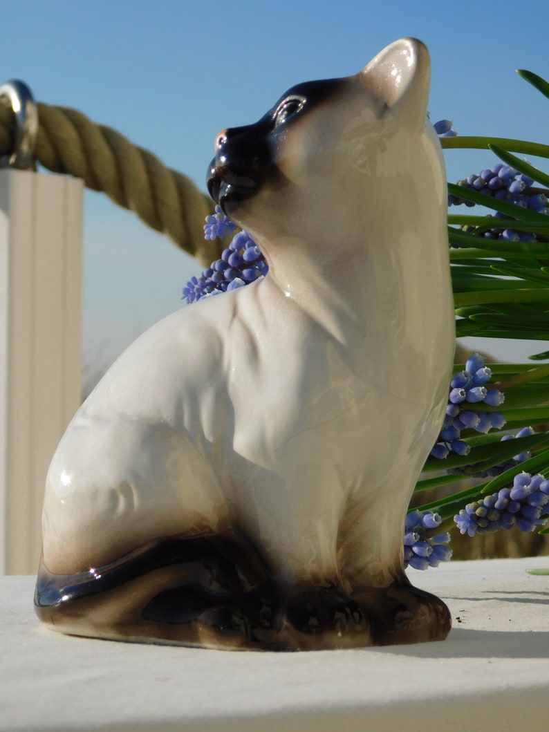 May include: A ceramic figurine of a Siamese cat sitting with its head turned to the side. The cat is white with black markings and has blue eyes. The figurine is sitting on a white surface with a bouquet of purple flowers in the background.