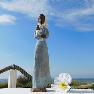 Peut inclure: Une figurine haute et élancée représentant un personnage vêtu d'une longue robe bleu clair texturée et d'un couvre-chef blanc, tenant un petit nourrisson en vêtements blancs. La sculpture présente une finition brun foncé sur le visage et les mains.