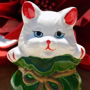 Könnte beinhalten: Weiße Keramik-Katzenfigur mit roten Ohren und grüner und brauner Kleidung. Die Katze trägt einen grünen und braunen Sack mit einem braunen Band darum.