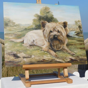 May include: A painting on an easel of a small, light brown dog with a fluffy coat, lying down in a grassy field. The painting is set against a backdrop of a blue sky and green trees.