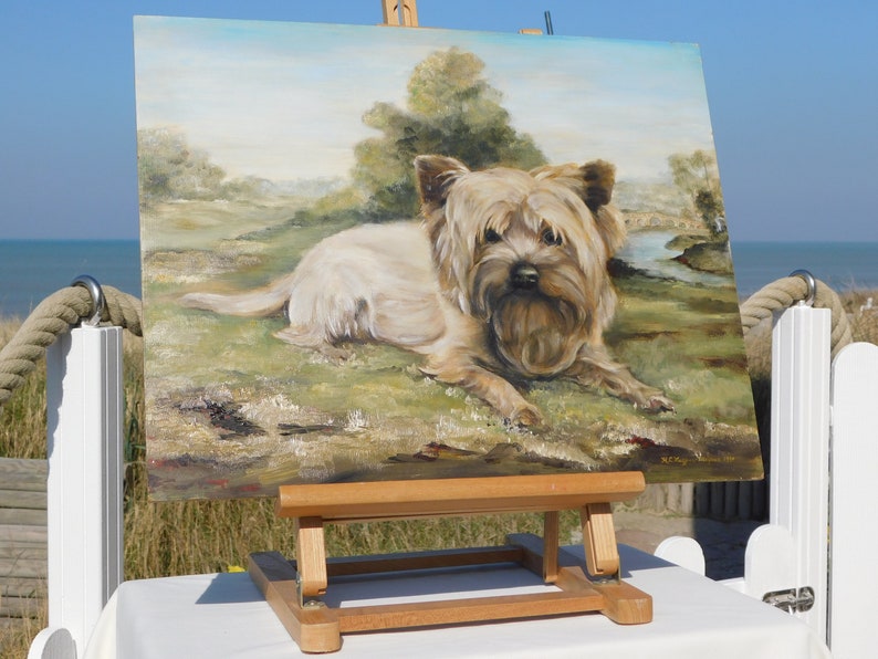 May include: A painting on an easel of a small, light brown dog with a fluffy coat, lying down in a grassy field. The painting is set against a backdrop of a blue sky and green trees.