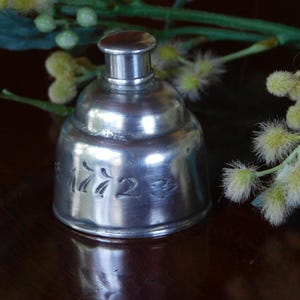 May include: A small, antique silver-colored bottle with a rounded body and a small, cylindrical top. The number "1723" is etched into the side of the bottle. The bottle is set against a dark wooden surface with yellow and green floral accents.