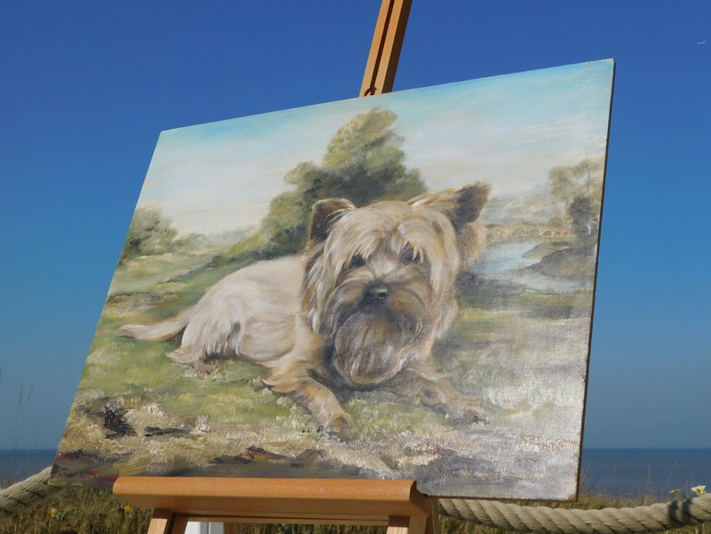 May include: A painting on canvas of a brown and white Yorkshire Terrier dog lying down in a grassy field. The dog is looking at the viewer with its tongue slightly sticking out. The background is a green field with a small stream and trees in the distance.
