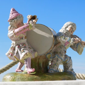Puede incluir: Una figurita de porcelana de dos payasos tocando música. Un payaso lleva un vestido rosa y blanco y sostiene un tambor. El otro payaso lleva un traje azul y blanco y toca el violín.
