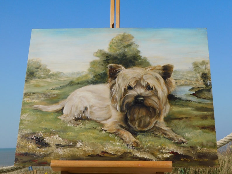 May include: A painting of a small, light brown dog with a fluffy coat, lying down in a grassy field. The dog is looking directly at the viewer. The painting is set against a backdrop of a blue sky and green trees.