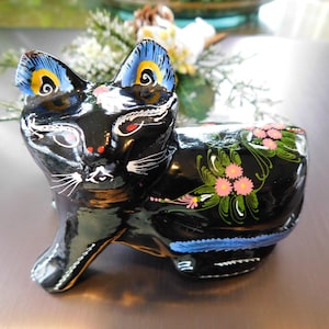 Puede incluir: Figura de gato de cerámica negra con orejas pintadas de azul y amarillo y detalles florales en la espalda. El gato tiene bigotes y ojos pintados de blanco.