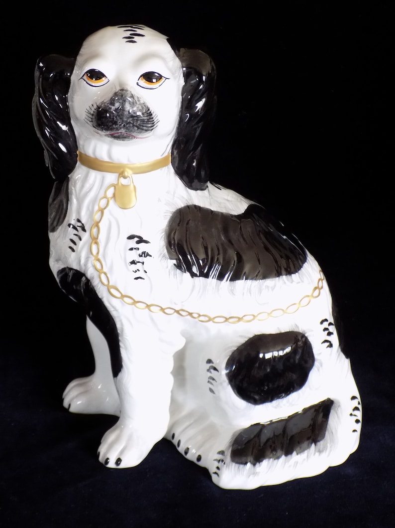 K&ouml;nnte beinhalten: Eine schwarz-wei&szlig;e Porzellanfigur eines Hundes mit goldenem Halsband und Kette. Der Hund sitzt mit dem Kopf zur Seite geneigt und die Ohren h&auml;ngen herunter. Die Figur ist mit realistischen Details bemalt, darunter das Fell, die Augen und die Nase des Hundes.