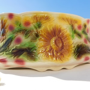 May include: A ceramic planter with a sunflower design. The planter is rectangular with a scalloped edge and is decorated with raised sunflowers in shades of yellow and brown, green leaves, and pink polka dots.