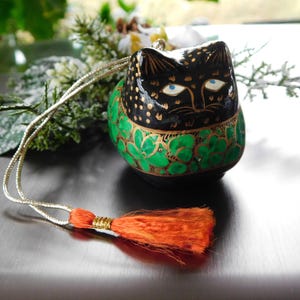 Puede incluir: Adorno en forma de gato negro con un diseño floral verde y dorado alrededor de la base. El gato tiene ojos azules y detalles dorados. Una borla naranja cuelga de un cordón dorado sujeto al adorno.
