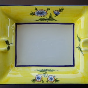 Puede incluir: Un cenicero de cerámica amarillo con un diseño floral azul y blanco. El cenicero tiene una forma rectangular con esquinas redondeadas y un borde elevado.
