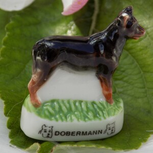 May include: A small ceramic figurine of a Doberman Pinscher dog. The figurine is brown and black with a white base and green grass. The base has the word "Doberman" written on it.